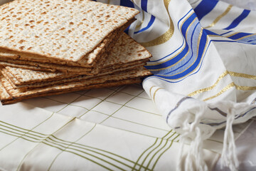 Stack of matzo, an unleavened flatbread, next to a Tallit, a Jewish prayer shawl. The matzo and Tallit are on a surface covered with a white and green grid patterned fabric. Jewish Passover holiday.