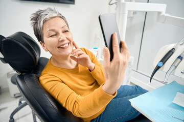 Woman sitting in medical chair and making selfie of her healthy and white teeth