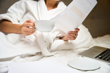 Cropped businesswoman with cup of tea or coffee watching document on bed i