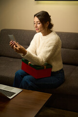 Woman watching smartphone on sofa and holding gift during Christmas or New Year