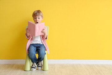 Cute little boy reading book while sitting on chair against yellow wall