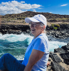 Senior woman in outdoors vacation at sea sitting on a rocky beach admiring waves splashing on the...