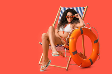 Beautiful young African-American female lifeguard with ring buoy sitting on deckchair against red...