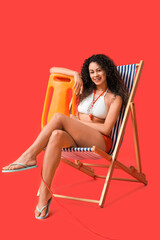 Beautiful young happy African-American female lifeguard with rescue buoy sitting on deckchair...