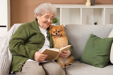Senior woman with cute Pomeranian dog reading book on sofa at home