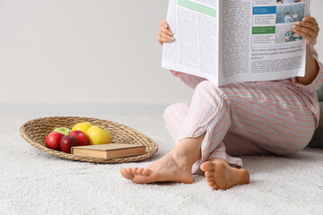 Barefoot woman in pajamas reading newspaper on white carpet at home