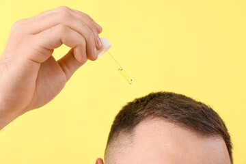 Young man using serum for hair growth on yellow background, closeup