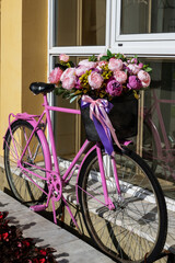 Pink vintage bicycle with a basket full of flowers next to a building with large windows. Street decoration in retro style - 791132069
