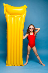 Obraz premium Swimming is fun. Happy little girl wearing red swim suit in sunglasses going to poll with yellow inflatable mattress, blue studio background. Summer vacation concept. Happy family