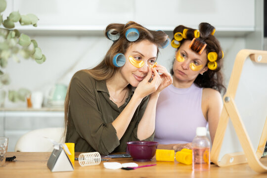 Two interested adult female friends in colorful curlers using golden moisturizing under-eye patches during daily self-care routine, sharing thoughts on skincare experiences in cozy home environment