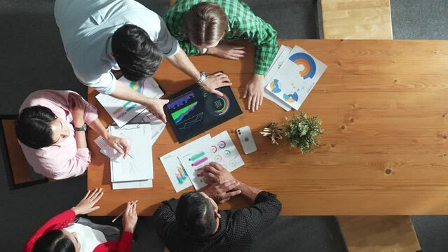 Top view of business man using tablet display increasing graph while diverse marketing people sharing idea or planing strategy at meeting table with stock market statistic scatter around. Convocation.