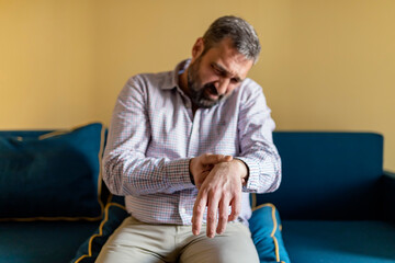 Mature Bearded Man Massaging His Painful Wrist. Man Suffering From Wrist Pain At Home, Sitting on...