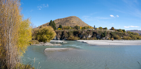 A panoramic view of the Shotover River, clear water flows rapidly over a shallow riverbed with...