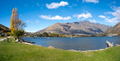 A panoramic view of Lake Wakatipu in Frankton, featuring a lush green grass lawn, a jetty, and the Remarkables mountain range in the background. Queenstown, New Zealand.