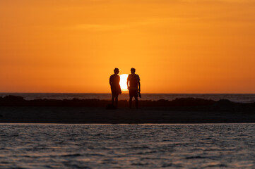 Fototapeta premium Man and woman staying and looking at sunset on ocean beach, orange sky, silhouettes of people on vacation