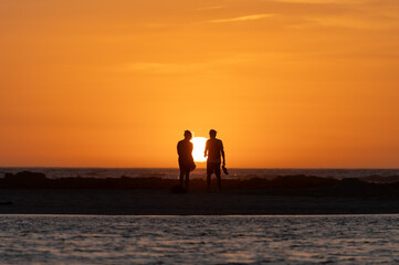 Fototapeta premium Man and woman staying and looking at sunset on ocean beach, orange sky, silhouettes of people on vacation