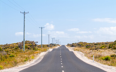 Fototapeta na wymiar A heavily undulating or bumpy road stretches into the distance with power lines and heat haze under the summer sky in Tasmania, Australia