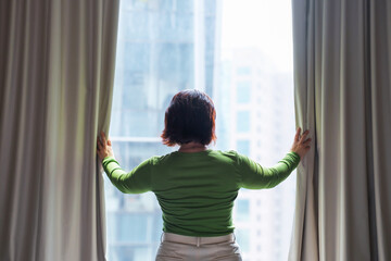 A woman opens curtains and looks out window.