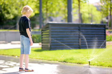 Preteen boy watching an automatic garden sprinkler during walks in a public park. Gardening equipment. Watering the grass on lawn on a sunny summer day