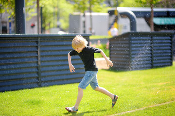 Funny little boy playing with lawn sprinkler in sunny city park. Elementary school child laughing, jumping and having fun with spray of water. - 791185802