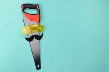 Man's face made of artificial mustache, safety glasses and hand saw on light blue background, top...