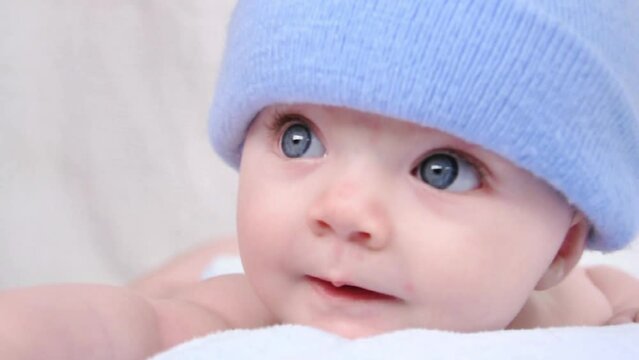 A little boy baby is hiding under a blue blanket with a hat on. He is staring at the camera and posing on his tummy.