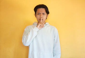 Portrait of serious Asian young man wearing a hoodie showing silence sign, promising to keep secret