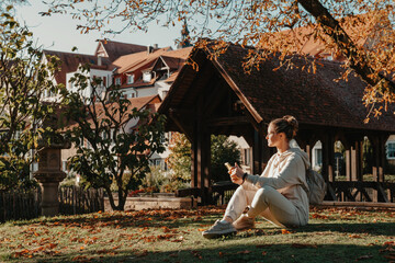 Obraz premium Young Fashionable Teenage Girl With Smartphone In Europian Park In Autumn Sitting At Smiling. Trendy Young Woman In Fall In Park Texting. Retouched, Vibrant Colors. Beautiful Blonde Teenage Girl