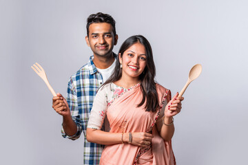 Asian indian young couple standing with kitchen utensils against plain background