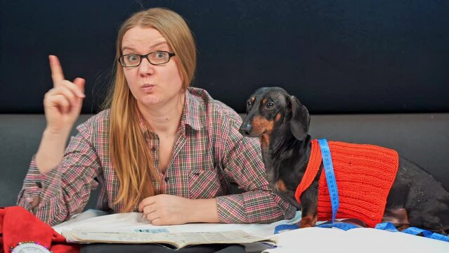 Young woman thoughtfully taps fingers on table near sitting dachshund. Female designer reflects on new ideas for animal clothes