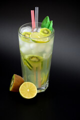 A tall faceted glass of refreshing tropical lemonade with ice and straws on a black background, next to slices of ripe lime and kiwi fruit.