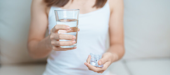 woman hand holding medicine painkiller pill and water glass on the sofa at home, taking for...