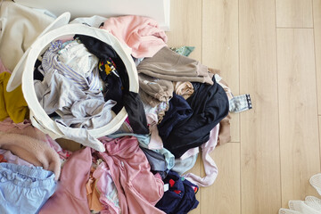 Clothes pile next to laundry basket filled with garments, on hardwood floor