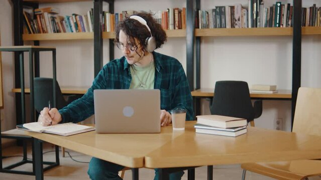Young guy college student sit at desk use pc laptop computer watching distance online learning seminar class. Remote university webinar having virtual classroom meeting in library. Man in headphones