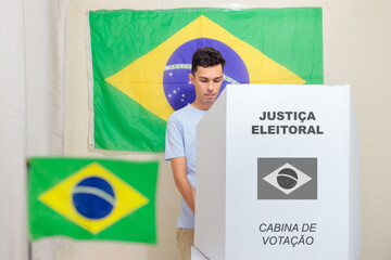 Jovem homem brasileiro escolhendo o seu candidato na urna eletrônica. Na parede, a bandeira do Brasil decora o local de votação. A imagem retrata as eleições brasileiras	