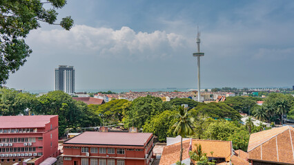 Panorama of the modern city. Multi-storey buildings, skyscrapers among lush green tropical vegetation. A tall TV tower against a blue sky and clouds. The ocean is far away. Malaysia. Malacca - obrazy, fototapety, plakaty