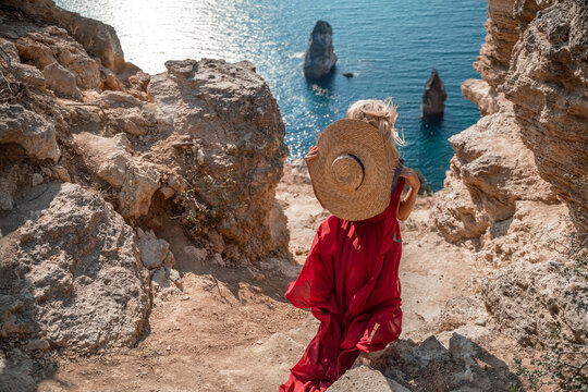 From behind, a woman is seen in a red flying dress fluttering in the wind. In a straw hat, walking down the stairs against the background of the sea with rocks and boats. The concept of romance