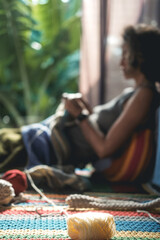In the foreground is a ball of woolen thread in the background out of focus is a young woman with a short haircut who sits on the floor and knits a blanket with a basket of tools standing next to her.