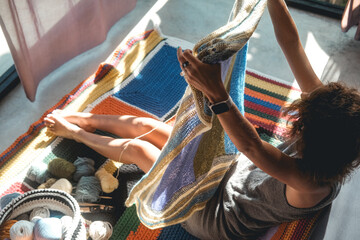 A young woman knits a multi colored and soft blanket from woolen threads. She raised it up to evaluate the quality of her work. She sits on the floor in her room, a quiet hobby.