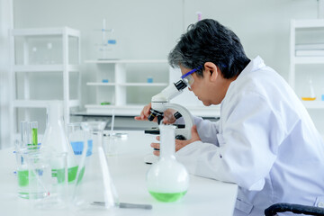 Focused Asian lab worker adjusting microscope, preparing slide, with test tubes and beakers...