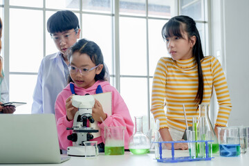 Students' learning in science. Two students, one observing through microscope, other looking on with curiosity, both in lab coats, amidst colorful lab glassware in bright classroom.