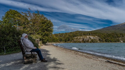 A man in a down jacket is sitting, resting on a log bench by the emerald lake. Smiling, looking into the distance. Green trees on the shore. Mountains against the blue sky. Argentina. Lago Roca. 