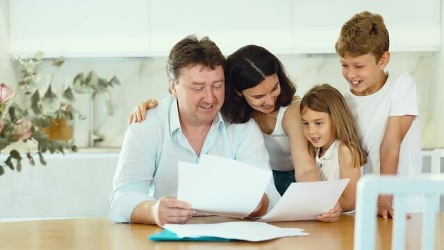 Positive husband is sitting at kitchen and reading document about career advancement at work, moneymaking financial report. Smiling wife and happy children hug successful dad