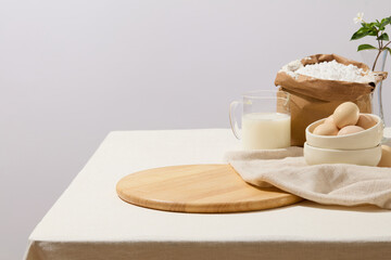 Fototapeta na wymiar An empty chopping board arranged on a table with a cup if milk, a paper bag containing flour, bowls of eggs and a towel. Food, bread or cake can be shown in the vacant space for advertising
