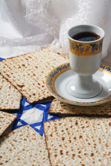 Jewish Passover celebration. A cup filled with red wine sits on a saucer, surrounded by matzah bread. The bread is flat and textured. All items rest on a cloth featuring the Star of David pattern