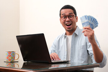 A man sitting in front of laptop showing wow expression at the camera while holding a lot of money