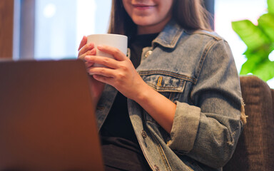 Closeup image of a young woman drinking coffee while working and looking at laptop computer - 791325270