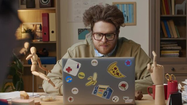 Medium over-shoulder shot of cooky male corporate worker with curly red hair, beard, in glasses sitting at desk, working with laptop at report, glancing over at pedantic colleague in suit and sighingM