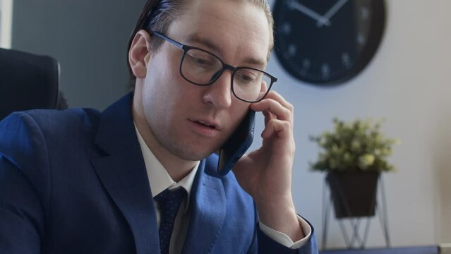 Medium close-up shot of narcissistic male office worker sitting at desk, having business conversation on mobile phone, looking at graphs, slicking back greased hair and straightening glassesMedium clo