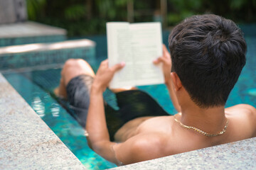 Asian man laying in the swimming pool reading a book in the summer on a sunny day. Relaxing in a...